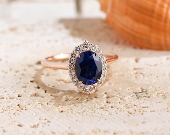 May include: A rose gold ring featuring a large, oval-cut deep blue sapphire center stone. The sapphire is surrounded by a halo of small, round, clear white diamonds set in a prong setting. The band is thin, smooth, and made of rose gold metal.