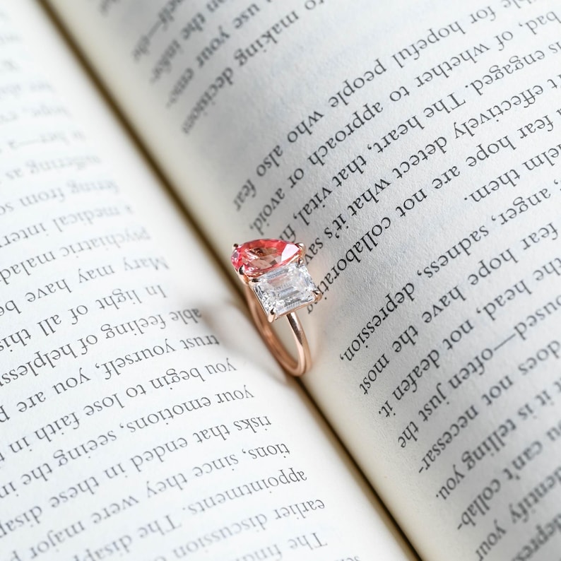 May include: A rose gold ring with a pink and clear gemstone setting. The ring is resting on an open book.