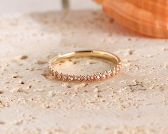 May include: A thin gold band ring featuring a continuous row of small, round, light pink gemstones set along the outer circumference. The metal has a polished finish, and the stones are uniform in size, creating a delicate, circular design.