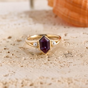 May include: A gold ring featuring a central hexagonal purple gemstone flanked by two small white pearls. The band is crafted from a polished yellow metal, and the setting holds the stones in a secure, symmetrical arrangement on the ring shank.