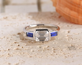 May include: A silver-toned ring featuring a large, rectangular, emerald-cut clear center stone flanked by two smaller, rectangular, deep blue sapphire-colored stones. The stones are set in a polished metal band with a clean, minimalist design.