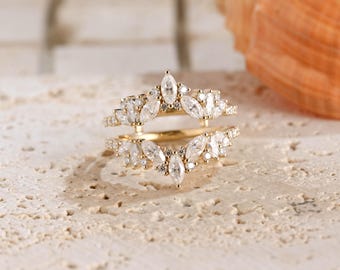 May include: A gold ring guard featuring two symmetrical bands connected at the base, adorned with multiple marquise-cut and round-cut clear stones arranged in a pointed, leaf-like pattern that frames a central opening for another ring.