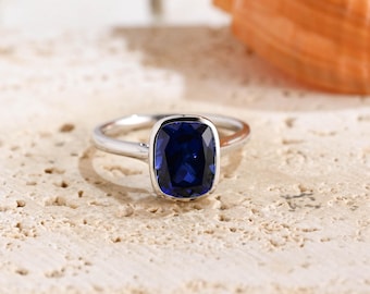 May include: A silver-toned ring featuring a single, large, cushion-cut deep blue gemstone set in a smooth bezel setting. The band is polished and metallic, showcasing a clean, simple design that highlights the vibrant color of the central stone.