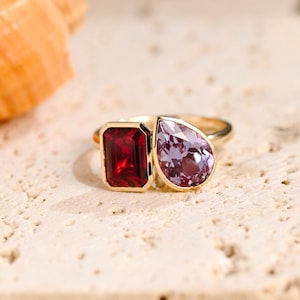 May include: A gold ring featuring two gemstones. The larger stone is a square emerald cut, deep red in colour. The smaller stone is a pear-shaped, light purple gemstone.