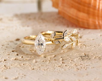 May include: A gold ring featuring a central marquise-cut clear gemstone and a band designed with multiple small leaf-shaped accents, each set with a tiny clear stone. The metal has a polished finish, and the design mimics a delicate vine.