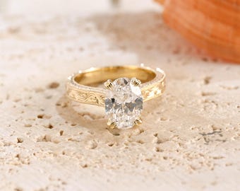 May include: A gold ring featuring a large, oval-cut clear gemstone held by four prongs. The band is crafted from yellow gold and displays an intricate, engraved scrollwork pattern along the sides, extending from the central stone setting.