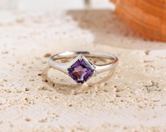May include: A silver-toned ring featuring a single square-cut purple gemstone set in a four-prong setting. The band is smooth and polished, tapering slightly toward the center where the faceted amethyst-colored stone is positioned.