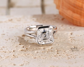 May include: A silver-toned ring featuring a square-cut, clear, faceted gemstone set in a bezel setting. The band has a split design that connects to the central stone setting, showcasing a polished metallic finish and a brilliant, reflective center.