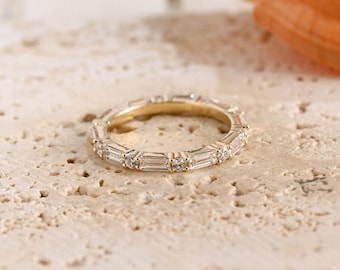 May include: A gold eternity band featuring an alternating pattern of baguette-cut and round-cut clear stones set along the entire circumference of the ring. The metal band has a polished finish and holds the stones in a continuous, repeating design.