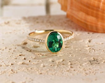 May include: A gold ring featuring a large, oval-cut, vibrant green gemstone set in a smooth bezel. The slender band is accented with a row of small, clear, sparkling stones embedded along both sides of the central green oval setting.