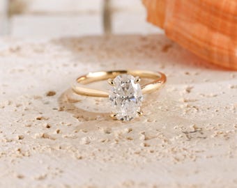 May include: A gold ring featuring a single oval-cut clear gemstone set in a four-prong setting. The band is smooth and polished, showcasing a classic design with a brilliant, faceted stone that reflects light from its center point.