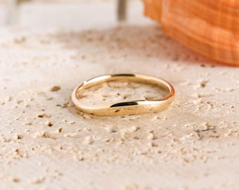 May include: A thin, polished gold ring with a smooth, continuous band. The ring features a subtle, gentle wave or curve along its circumference, creating a fluid and rounded shape. The metal surface is reflective and uniform in its golden tone.