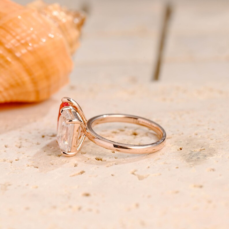 May include: A rose gold engagement ring with a large, square-shaped, clear gemstone. The ring has two smaller, red gemstones on either side of the main stone.