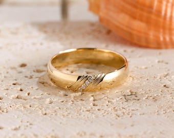 May include: A gold band ring featuring a polished finish and a diagonal section with three small, clear, round-cut stones set in a row. The band has a smooth, rounded profile and a bright, reflective metallic surface throughout its circular structure.