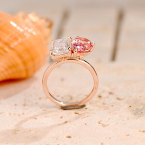 May include: A rose gold ring with two gemstones. The larger gemstone is a square-shaped clear stone, and the smaller gemstone is a pear-shaped pink stone.
