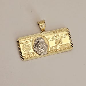 May include: Gold-toned pendant in the shape of a $100 bill. The pendant features a detailed depiction of the bill's design, including the portrait of Benjamin Franklin and the text "The United States of America".