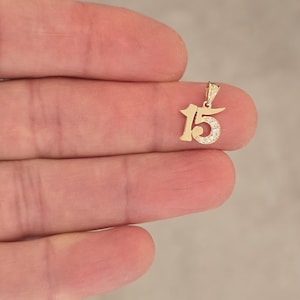 May include: A gold pendant shaped as the number 15, featuring a polished finish on the number one and a curved section on the number five that is inlaid with small, clear, sparkling stones, complete with a small triangular bail at the top.