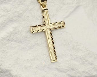 May include: A gold cross pendant featuring a textured, diamond-cut finish on the surface. The pendant includes a polished gold bail at the top for attachment to a chain, showcasing a detailed, reflective pattern across the cross arms.