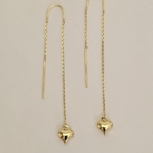 May include: A pair of gold-colored threader earrings. Each earring features a curved top, a delicate chain, and a teardrop-shaped charm. The earrings are displayed against a plain, light background.
