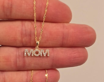 May include: A gold-toned necklace featuring a pendant that spells out the word MOM in capital letters. The pendant is encrusted with small, clear, sparkling stones. The necklace is attached to a delicate, twisted gold-toned chain.