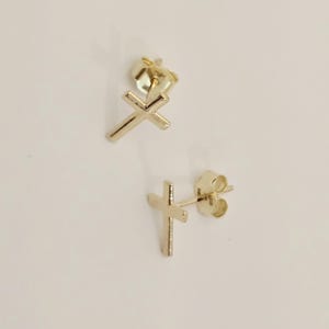 May include: A pair of gold-colored cross stud earrings. The earrings are small, with a simple cross design. The posts and backs are visible, suggesting a secure fastening. The earrings are set against a plain, light background.