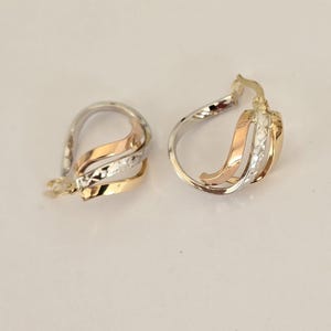 May include: A pair of hoop earrings crafted from tri-color gold, featuring intertwined ribbons of yellow, rose, and white gold. The white gold sections display a textured, diamond-cut finish, while the yellow and rose gold bands are smooth.