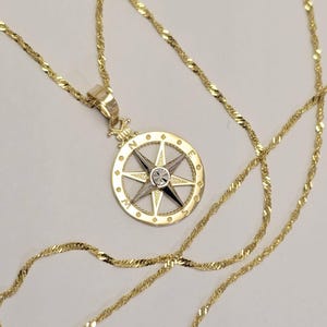 May include: A gold compass pendant necklace with a diamond-shaped stone in the center. The compass is surrounded by a circle with the letters N, E, S, and W.