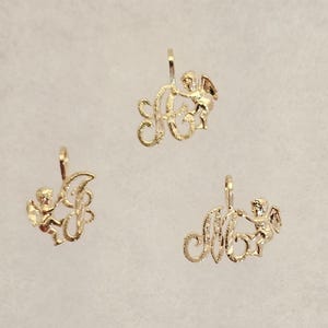 May include: Three gold-toned metal charms, each featuring a textured script letter intertwined with a small cherub figure. Each charm includes a small loop at the top for attachment, showcasing a polished finish with intricate detailing on the figures.