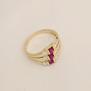 May include: A gold ring featuring a cluster of three rectangular, deep red gemstones, accented by smaller, round, clear stones. The ring has a triple-band design, with the gemstones set in a central, raised section. The ring is displayed on a light-colored surface.