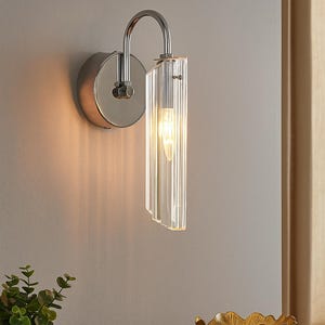 Op de afbeelding: Een moderne wandlamp met een chromen afwerking en een heldere, geribbelde glazen kap. De lamp is gemonteerd op een lichtgrijze muur, met een warme gloed. Een gouden kom en een kleine plant staan op een houten oppervlak.