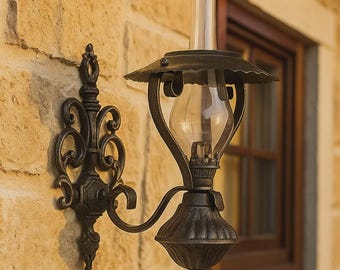 May include: An ornate, dark bronze wall-mounted lamp with a clear glass globe and a decorative bracket. The lamp is attached to a light-colored, textured stone wall. A wooden window frame is visible in the background.