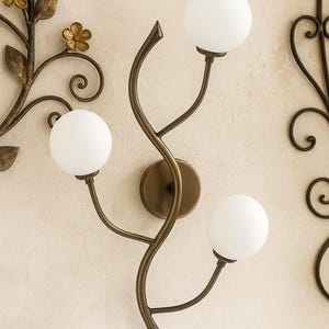 Può includere: Applique da parete decorativa con tre luci a globo bianche. L'apparecchio ha una finitura bronzo con un design curvo a forma di vite. Sono visibili ulteriori accenti floreali in bronzo.