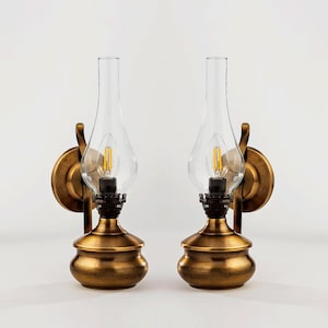 May include: Two antique-style wall sconces with a brass finish. Each sconce features a clear glass globe, a decorative brass base, and a small lightbulb. The sconces are mounted on a round brass plate with a curved arm.