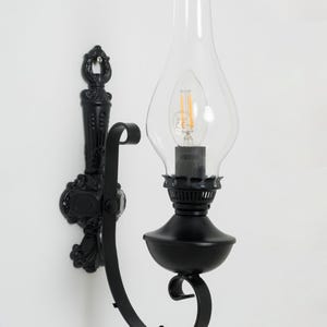 Puede incluir: Aplique de pared negro con una pantalla de cristal transparente y un diseño ornamentado. El aplique tiene una base de metal negro con un brazo curvado y una bombilla dentro de la pantalla de cristal. Montado en una pared blanca.