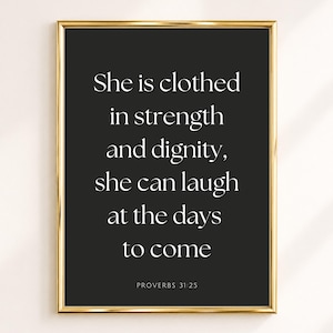 Op de afbeelding: Een rechthoekige wandkunstprint in een dunne gouden lijst met een zwarte achtergrond en witte schreefletters met de tekst: She is clothed in strength and dignity, she can laugh at the days to come. Onder de tekst staat de vermelding PROVERBS 31:25.