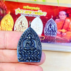 Könnte beinhalten: Dunkelblauer Anhänger mit einer sitzenden Buddha-Figur in einem bogenförmigen Rahmen. Der Anhänger wird in einer Hand gehalten, mit anderen ähnlichen Anhängern und einem Bild eines Mönchs im Hintergrund. Der Anhänger ist wahrscheinlich ein religiöses Objekt.