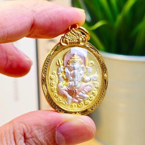 May include: An oval gold pendant with a detailed image of a deity with an elephant head and multiple arms. The deity is in silver and rose gold tones, set against a gold background. The pendant is held by a hand.