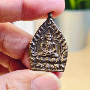 Könnte beinhalten: Ein bronzefarbener Anhänger mit einer sitzenden Buddha-Figur in der Lotusposition. Der Anhänger hat einen verzierten, blattartigen Rand und eine kleine Schlaufe oben zum Aufhängen. Der Buddha wird mit einem ruhigen Ausdruck dargestellt.
