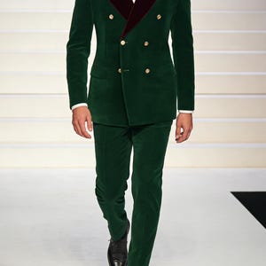 Könnte beinhalten: Ein grüner Samtanzug mit bordeauxfarbenem Revers und goldenen Knöpfen. Die doppelreihige Jacke wird mit einer passenden Hose kombiniert. Eine Fliege und ein Einstecktuch vervollständigen das formelle Ensemble. Schwarze Schuhe.