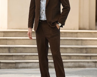 May include: A dark brown suit consisting of a single-breasted blazer with notched lapels and matching trousers. The suit is worn over a light-colored button-down shirt featuring a blue and grey botanical print pattern and dark brown leather shoes.