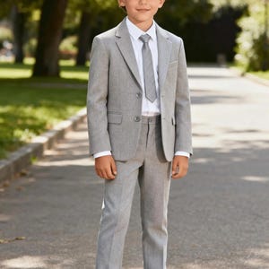 Op de afbeelding: Een lichtgrijs pak, bestaande uit een blazer, broek en stropdas, gecombineerd met een wit overhemd en witte sneakers. Het pak heeft een klassieke, op maat gemaakte pasvorm. De jongen staat op een geplaveide weg.