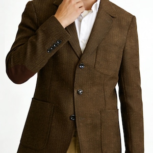 May include: A brown textured blazer featuring a three-button front closure, a chest pocket, two lower patch pockets, and dark brown elbow patches. The blazer is worn over a white collared shirt and tan trousers, showcasing a classic tailored design.