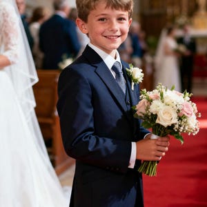 Puede incluir: Un joven con un traje azul marino, camisa blanca y corbata estampada, sosteniendo un ramo de flores blancas y rosas. La persona sonríe, con una rosa blanca prendida en la solapa. El fondo muestra una iglesia con una alfombra roja.