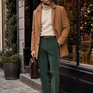 Op de afbeelding: Een camelkleurige wollen jas gedragen over een crèmekleurige coltrui en een donkergroene corduroy broek. De outfit bevat een bruine leren riem, bruine leren loafers en een bruine leren aktetas die in één hand wordt vastgehouden.