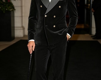 May include: A black velvet double-breasted tuxedo jacket with satin peak lapels, gold buttons, and a matching pair of trousers. The ensemble includes a white dress shirt, a dark green bow tie, a pocket square, and a black walking cane with a gold tip.