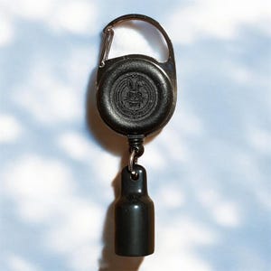 Könnte beinhalten: Eine schwarze, einziehbare Ausweishalterung mit einem Karabinerhaken aus Metall. Die Rolle hat ein kreisförmiges Design mit einem geprägten Emblem. Ein schwarzer, zylindrischer Anhänger hängt an der Rolle. Der Hintergrund ist hellblau mit weißen wolkenartigen Formen.