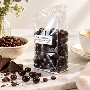 Puede incluir: Una bolsa de plástico transparente llena de granos de café cubiertos de chocolate negro. Una etiqueta rectangular blanca en la parte frontal dice "Dark Chocolate Coffee Beans 150g". Varios granos sueltos y trozos de chocolate están esparcidos.