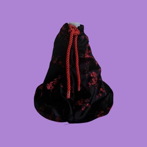 Puede incluir: Una bolsa de terciopelo negro con cierre de cordón rojo. La bolsa tiene un patrón floral y está decorada con borlas rojas.