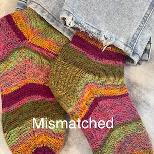 May include: A pair of hand-knitted ankle socks featuring a variegated yarn pattern in shades of magenta, olive green, orange, and yellow. The socks have a ribbed cuff and the word Mismatched is printed in white text across the center of the image.