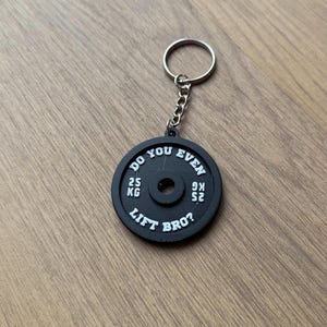 Könnte beinhalten: Schwarzer Gewichtsscheiben-Schlüsselanhänger mit weißem Text, der "Do you even lift bro?" und "25 kg" und "52.9 lb" lautet.